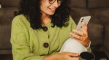 Девушка в свитере с чашкой кофе улыбается, листая ленту в телефоне