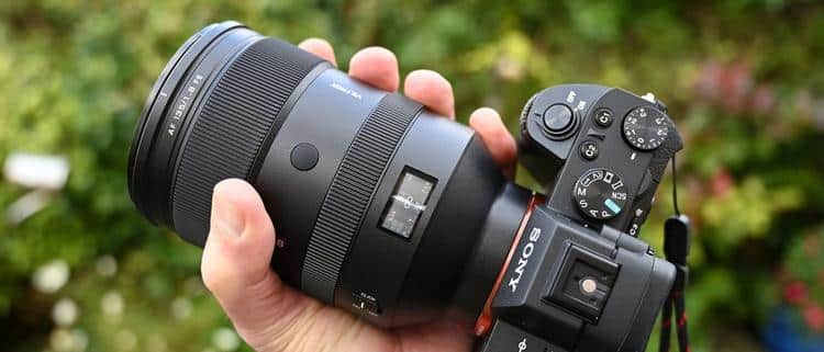 Мужская рука держит на улице объектив Viltrox AF 135mm f/1.8 LAB FE на камере Sony Alpha с активным цветным экраном дистанций и кольцом диафрагмы на фоне листвы.