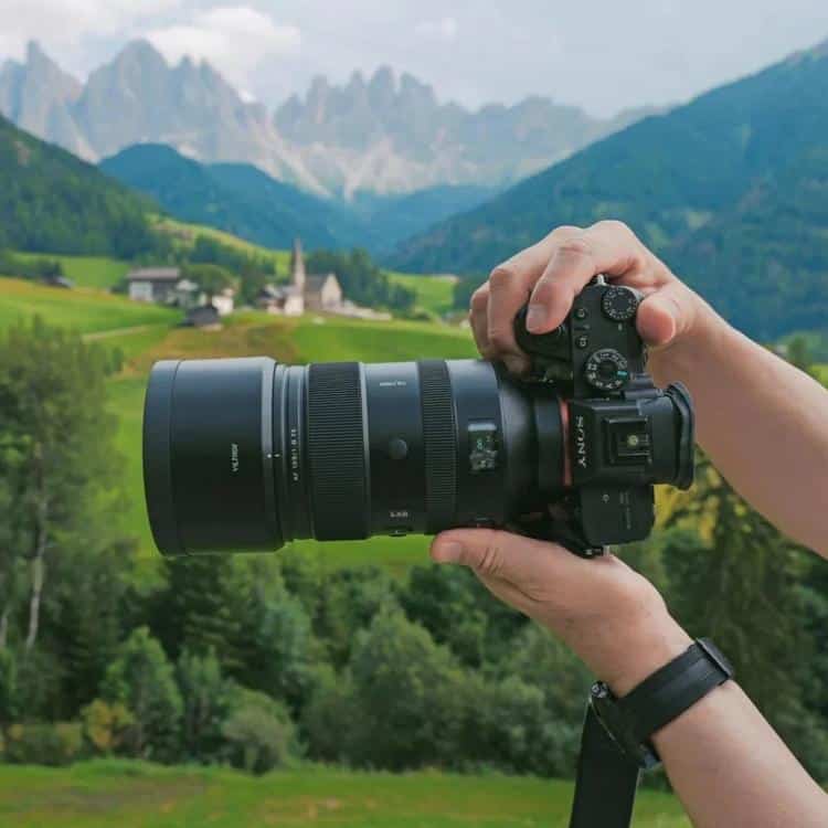 Фотограф держит двумя руками камеру Sony Alpha с длиннофокусным телеобъективом Viltrox AF 135mm f/1.8 LAB и надетой блендой во время съемки на фоне горной панорамы Доломитовых Альп.