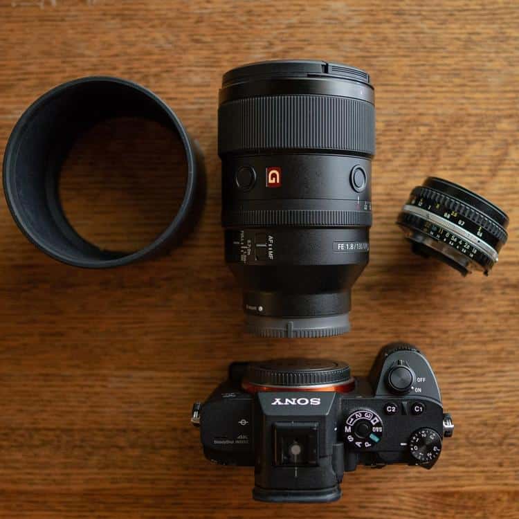 Разобранный комплект на деревянном столе: телеобъектив Sony FE 135mm f/1.8 GM с переключателями лимитера фокуса, бленда, тушка камеры Sony Alpha без объектива и компактный фикс 50 мм.