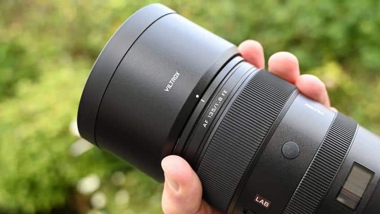 Рука держит переднюю осадку объектива Viltrox AF 135mm f/1.8 LAB с надетой глубокой цилиндрической блендой, широким кольцом фокусировки и красным логотипом серии LAB на открытом воздухе.