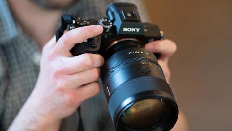 Руки фотографа в клетчатой рубашке держат установленный на камеру Sony Alpha объектив Sony FE 135mm f/1.8 GM с рифлеными кольцами управления и передней линзой в рассеянном комнатном свете.